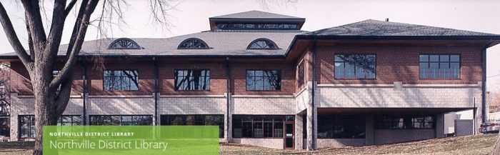 A modern brick building with large windows, featuring a sloped roof and decorative arches, surrounded by trees.